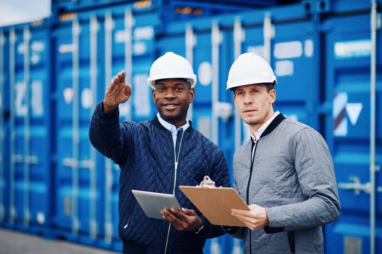 Two men in white hard hats holding clipboards.